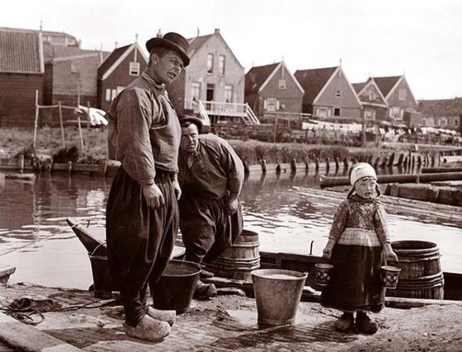 Два рыбака в мешковатых штанах деревянных башмаках стоят на причале вместе с маленькой девочкой держащей маленькие ведерца Маркен Нидерланды